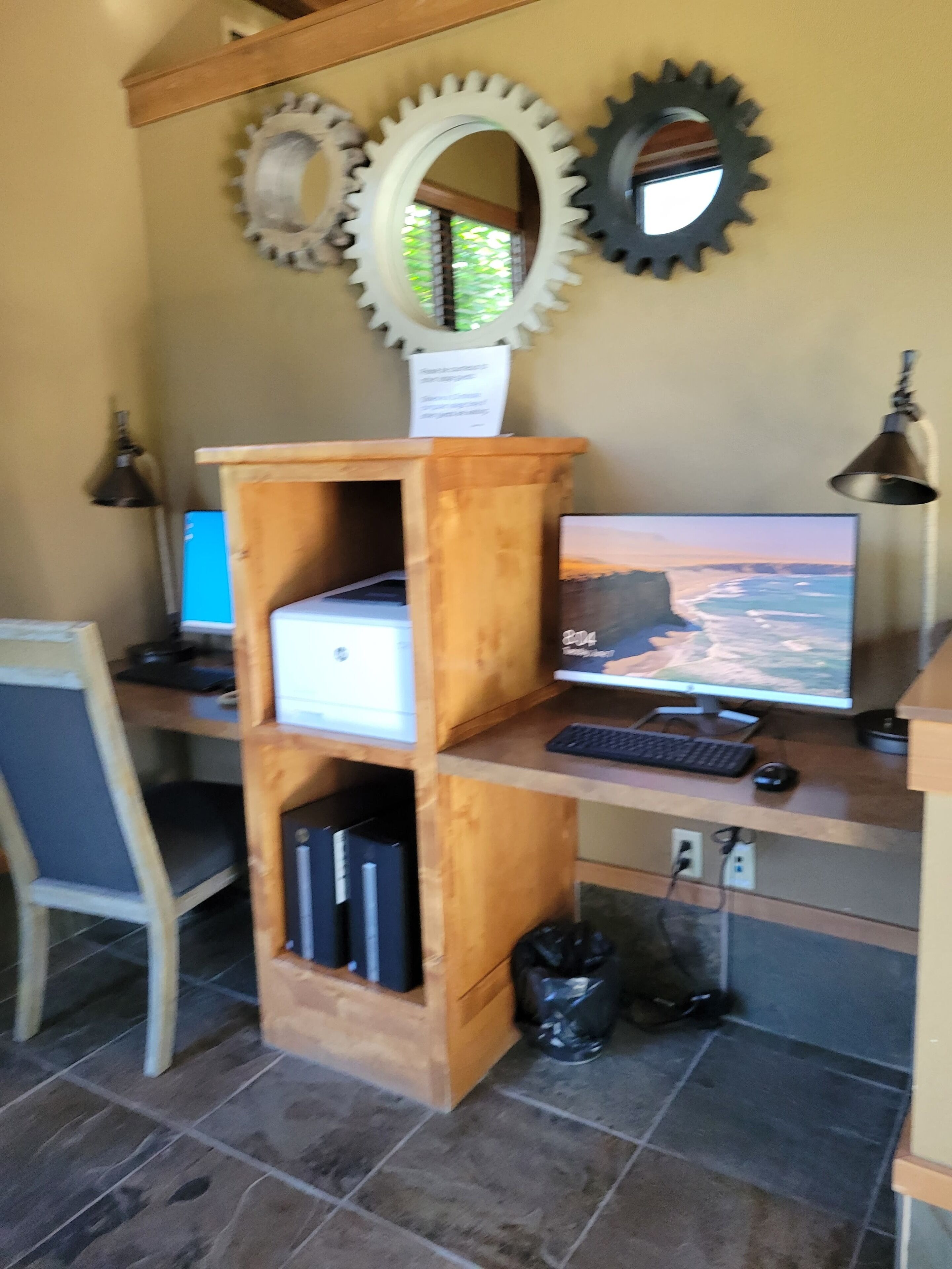 A wooden desk with a computer setup, including a monitor, keyboard, and mouse. There are two wall-mounted mirrors and a lamp.