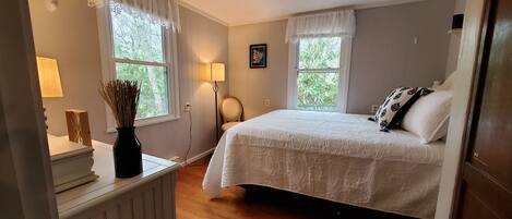 A bedroom with a bed, two windows with curtains, a bedside lamp, a chair, and a radiator.