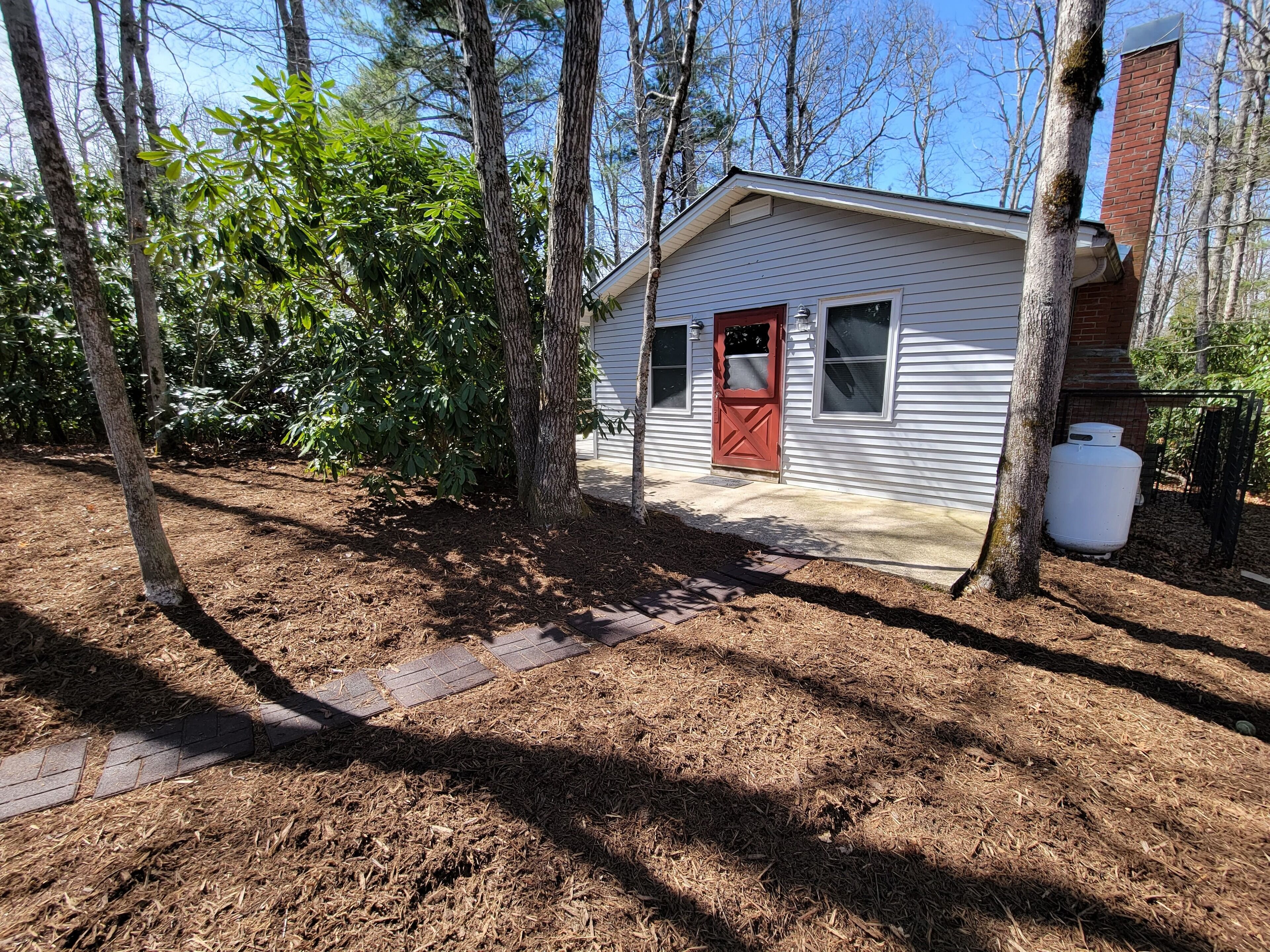 A small, single-story house with a red door and white siding, surrounded by trees and a yard with mulch.