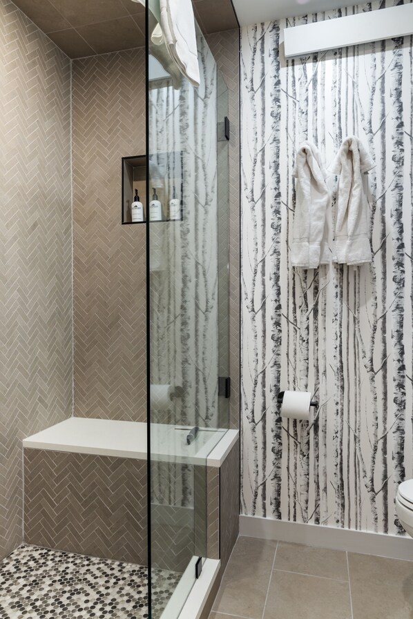 A modern bathroom with a herringbone tile pattern, a glass shower enclosure, and a toilet with a black and white tree wallpaper. - Outpost: Pines 322 - Central Wilson Location (Wilson)