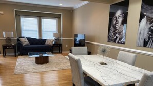 A living room with a dark blue sofa, a coffee table, and a dining table with chairs. There is a television on a stand and artwork on the walls. - An Eclectic Home for the World Traveler (Washington)