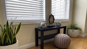 A room with a wooden floor, a white potted plant, a dark wooden side table with decorative items, and a round knitted pouf. - An Eclectic Home for the World Traveler (Washington)