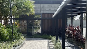 A paved pathway leading to a covered area with modern architecture, surrounded by various plants and greenery.