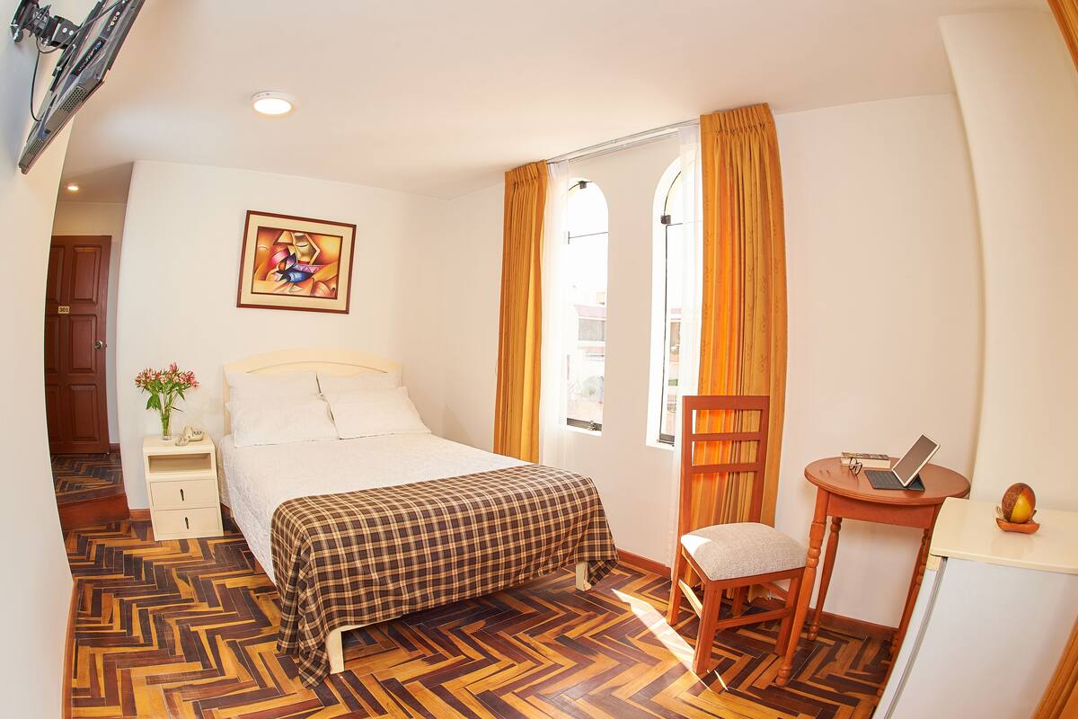 A bedroom with a bed, a chair, a desk with a laptop, a painting on the wall, and a window with curtains.
