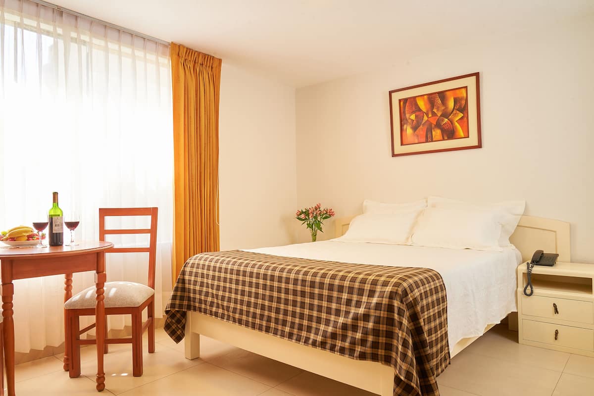A hotel room with a bed, a bedside table with a phone, a chair, a table with a bottle and glasses, and a painting on the wall.