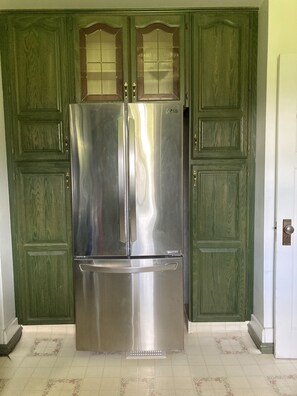 Uma geladeira de aço inoxidável com porta dupla e gaveta de freezer, posicionada entre dois armários de madeira verde com portas de vidro.