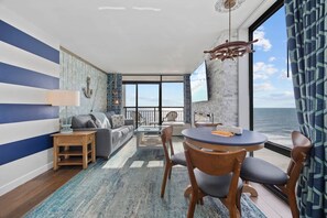 Ein Zimmer mit großem Fenster, einem runden Tisch mit Stühlen, einer Couch, einem an der Wand befestigten Fernseher und einem gemusterten Teppich.