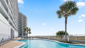 A swimming pool with a curved edge, surrounded by lounge chairs and a palm tree. - Lighthouse 703 "Sea Haven" Beachfront condo with huge balcony! (Gulf Shores)