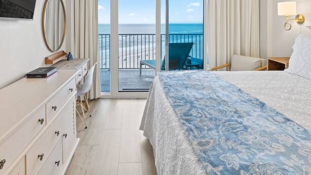 A hotel room with a bed, a desk, a chair, a TV, and a balcony with a view of the beach. - Lighthouse 703 "Sea Haven" Beachfront condo with huge balcony! (Gulf Shores)