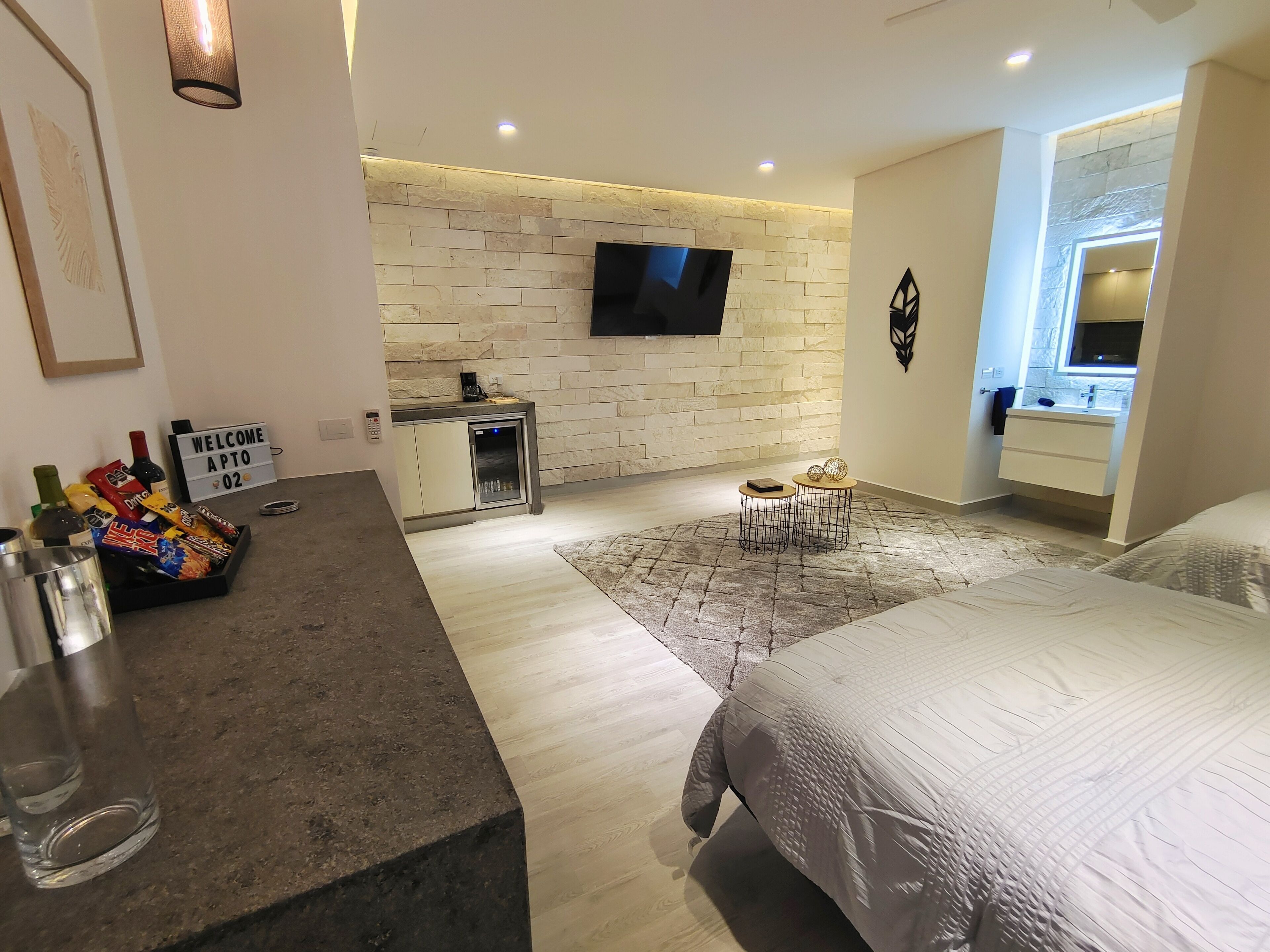 A modern hotel room with a stone wall, a flat-screen TV, a minibar, a small round table with two chairs, and a bed with a patterned cover.