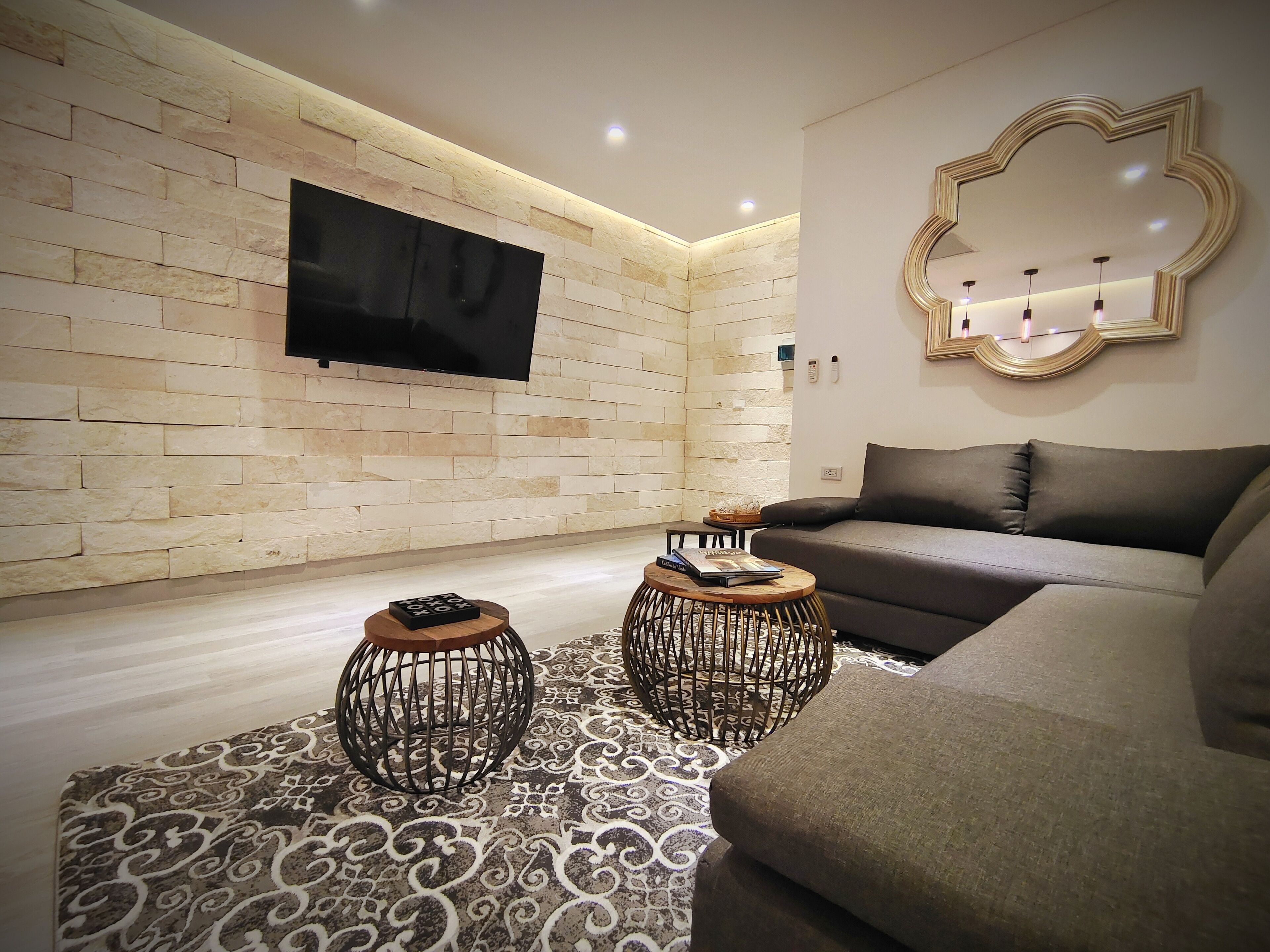 Una sala moderna con una pared de piedra, un televisor de pantalla plana, un sofá, dos mesas auxiliares decorativas y una alfombra estampada.