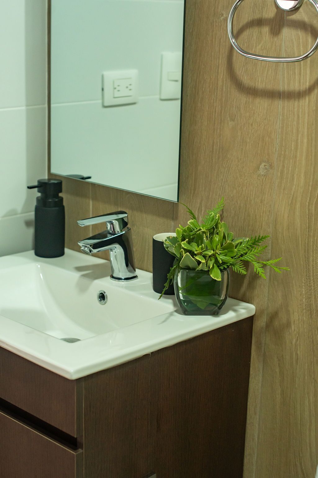 Ein Badezimmer mit einem weißen Waschbecken, einem modernen Wasserhahn, einem schwarzen Seifenspender und einer Zimmerpflanze in einer Glasvase.