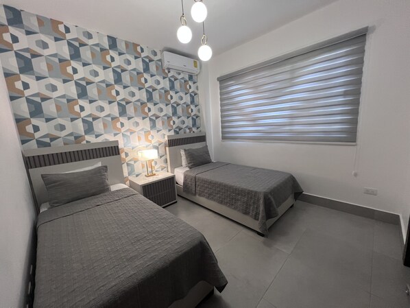 A bedroom with two beds, a geometric-patterned wall, an air conditioning unit, a bedside lamp, and a window with blinds.