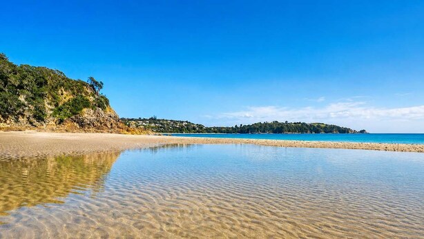 A beach with clear shallow waters, a sandy shore, and a distant hill with vegetation.