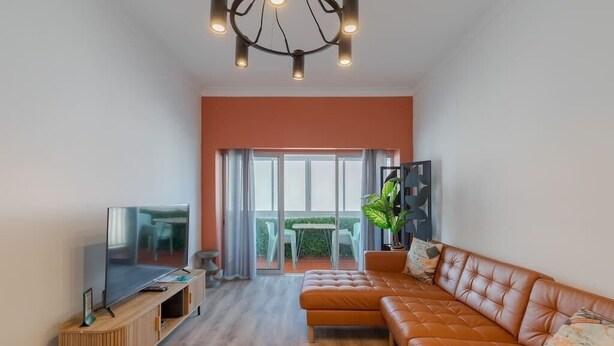 Un salon moderne avec un grand canapé d’angle, un téléviseur à écran plat posé sur une console en bois et une grande fenêtre donnant sur un balcon.