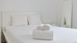 Ein ordentlich bezogenes Bett mit weißen Leinen und zusammengerollten Handtüchern, ein weißer Nachttisch und ein großer Spiegel.