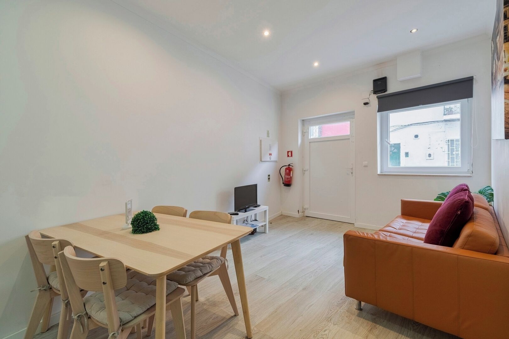 A modern living room with a wooden dining table, two chairs, a sofa with a cushion, a television, a fire extinguisher, and a window with a view of buildings.