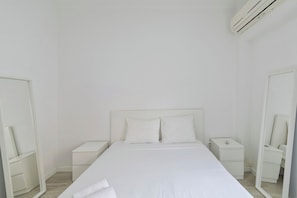 A minimalist white room with a bed, bedside tables, a mirror, and an air conditioning unit. - Gama Apartment (Setúbal)