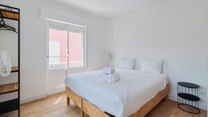 Een slaapkamer met een houten bed, witte beddengoed, een nachtkastje, een hanglamp en een raam met uitzicht op een roze gebouw.