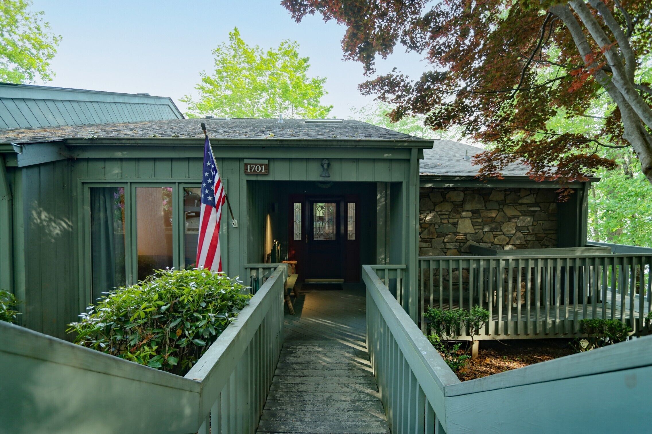 A small, green building with a stone wall and a wooden porch. An American flag is displayed outside.