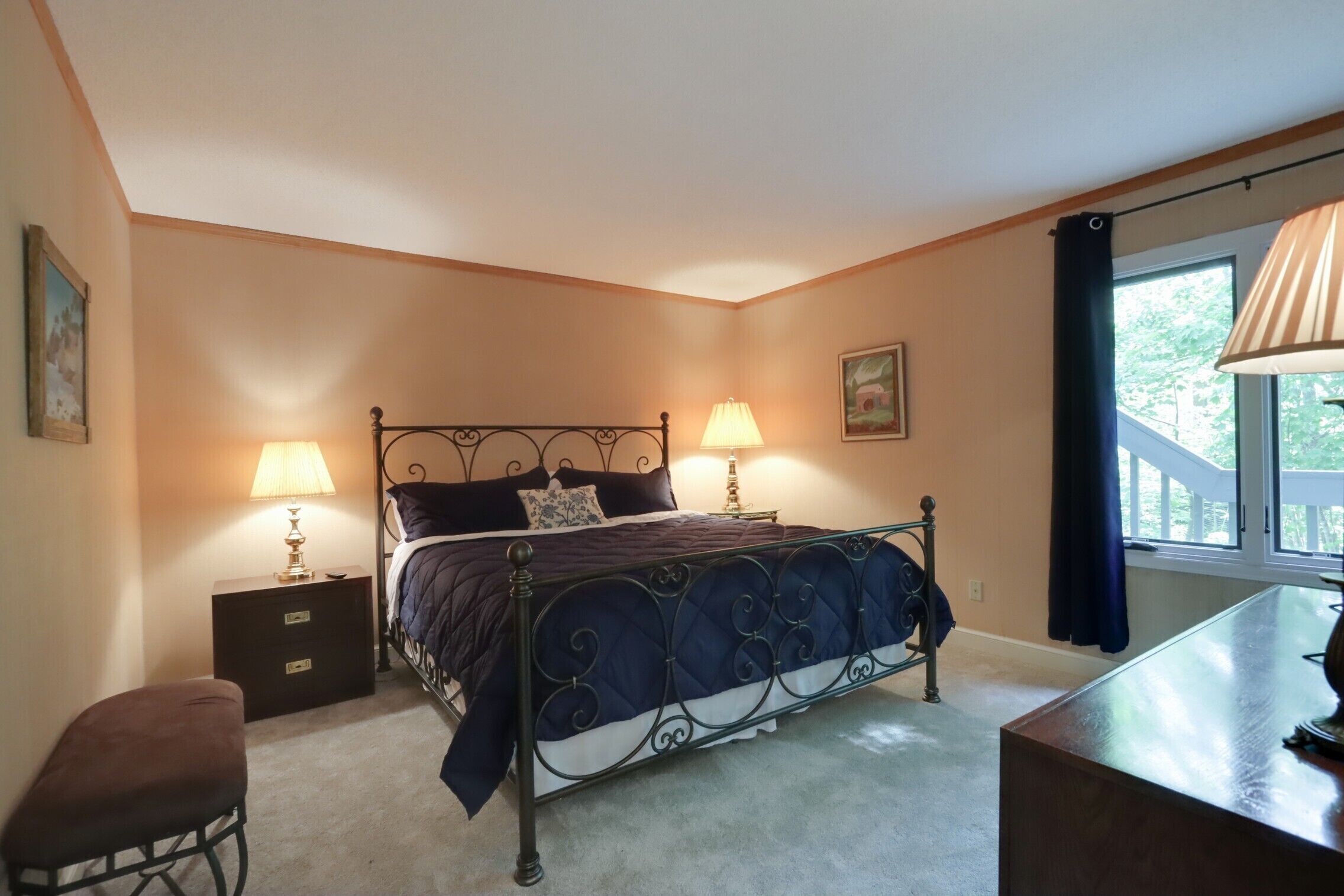 A bedroom with a bed, two bedside tables, a bench, a window with curtains, and a lamp.