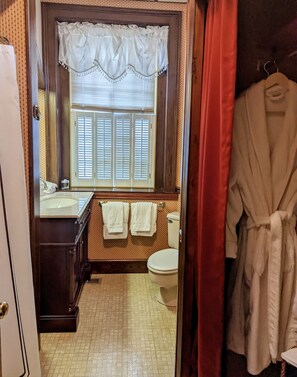 A bathroom with a toilet, sink, and window with shutters. There are towels hanging on the wall and a robe hanging on a rack. - Chanceford Hall Bed & Breakfast (Snow Hill)
