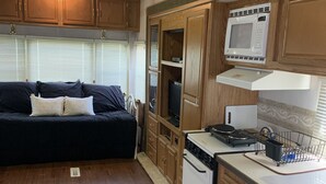 A compact RV kitchen with wooden cabinets, a microwave, stove, and a small dining area with a couch.