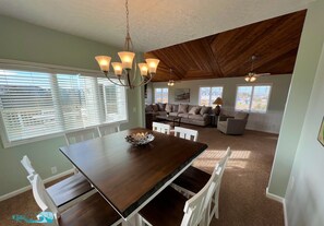 Un comedor con una mesa de madera y sillas blancas, una araña y vistas a una sala de estar con un sofá y una lámpara.
