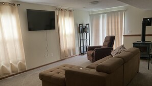 Ein Wohnzimmer mit einem braunen Ecksofa, einem flachbildfernseher an der Wand, einem Kamin und einem Fenster mit Jalousien.