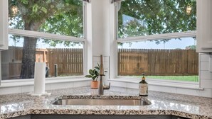 Eine Küche mit Granit Arbeitsplatten, einer Spüle aus Edelstahl und einem Fenster mit Blick auf einen Garten mit Holzzaun und Bäumen.