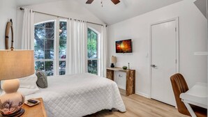 Ein Schlafzimmer mit einem Bett, einem Schreibtisch, einem Stuhl, einem Deckenventilator, einem Gemälde und großen Fenstern.