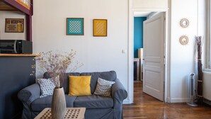 Uma sala de estar com um sofá cinzento, uma almofada amarela e um vaso com flores secas. Há duas obras de arte emolduradas na parede e um micro-ondas numa prateleira.