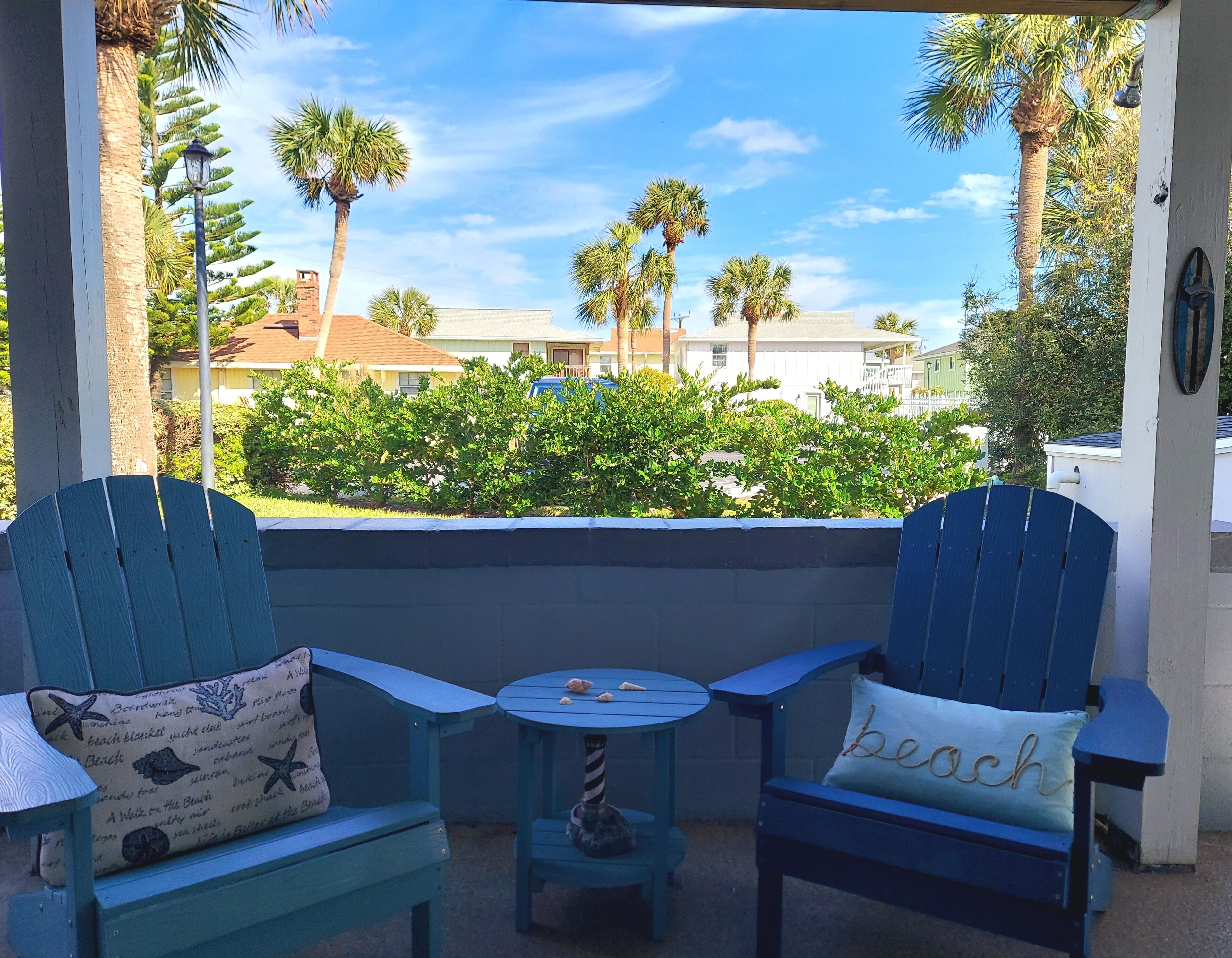 A covered outdoor seating area with two blue Adirondack chairs and a small round table, overlooking a residential neighborhood with palm trees and houses.