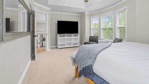 A bedroom with a large bed, a TV on a white stand, a chair, and a window with blinds. - Spacious 5-Bed Home with Pool in Overland Par! 27 (Overland Park)
