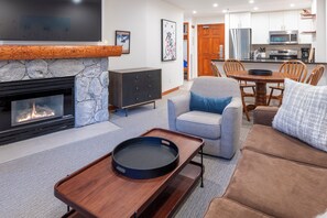 A modern living room with a stone fireplace, a flat-screen TV, a dining area with a wooden table and chairs, and a kitchen with stainless steel appliances.