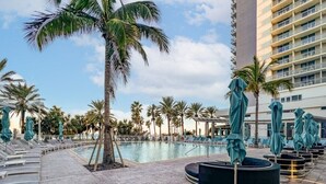 A poolside area with lounge chairs and umbrellas, surrounded by palm trees and a tall building in the background. - Wyndham Clearwater Beach Resort - 4 Bedroom (Clearwater Beach)