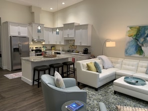 A modern kitchen with white cabinets, stainless steel appliances, and pendant lighting. There is a white sofa with a blue and white patterned rug in the living area.