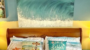 A bedroom with a bed featuring beach-themed pillows, a large abstract painting of waves, and a wooden headboard.