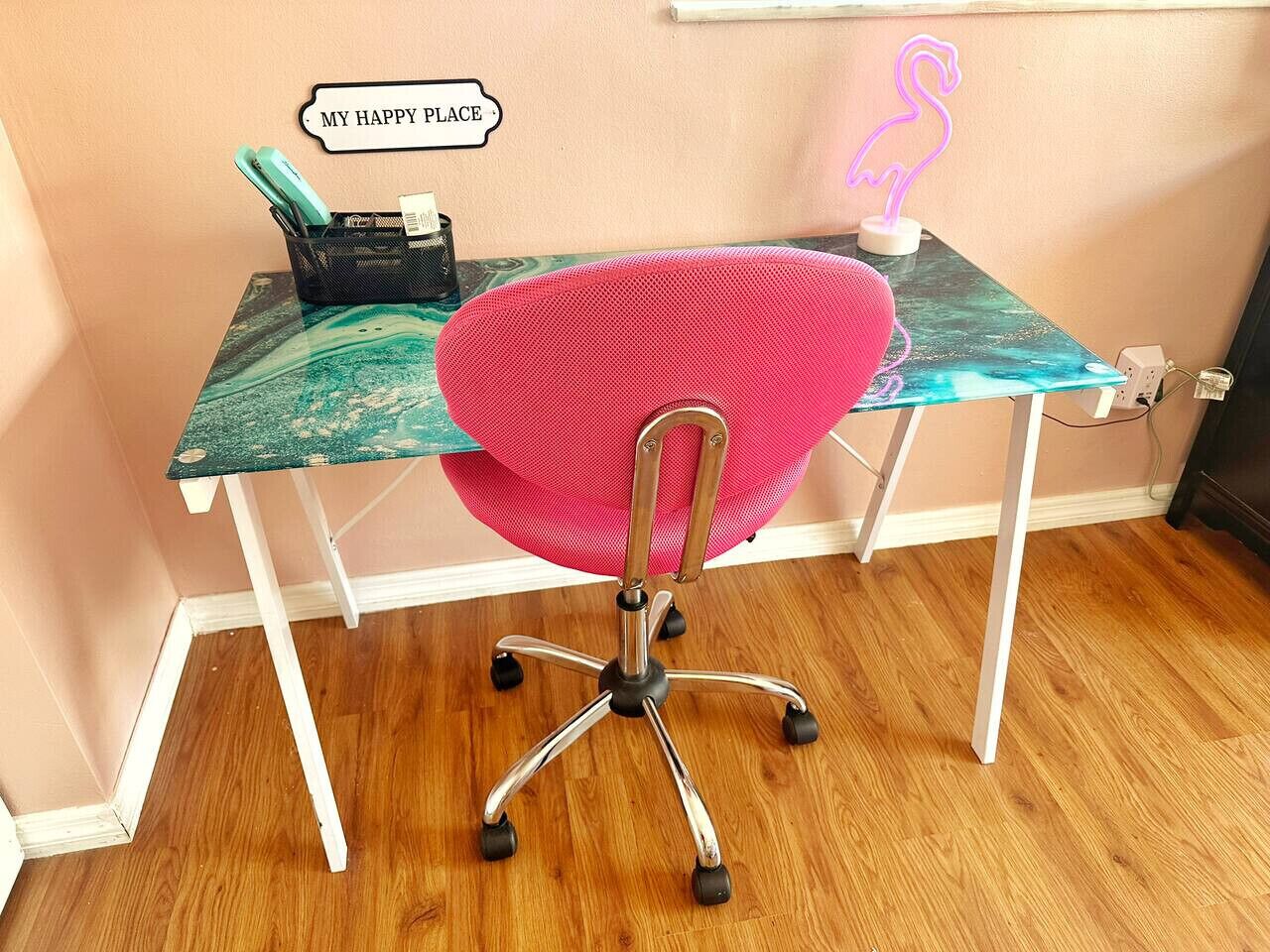 Ein rosa Bürostuhl mit Metallgestell und Rollen, ein Schreibtisch mit Türkis- und Grüntönungen im Marmor-Muster, ein schwarzer Netzkorb mit Gegenständen und ein neonfarbenes Flamingoschild an der Wand.