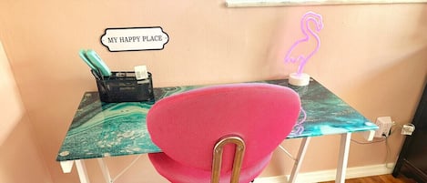 Ein rosa Bürostuhl mit Metallgestell und Rollen, ein Schreibtisch mit Türkis- und Grüntönungen im Marmor-Muster, ein schwarzer Netzkorb mit Gegenständen und ein neonfarbenes Flamingoschild an der Wand.