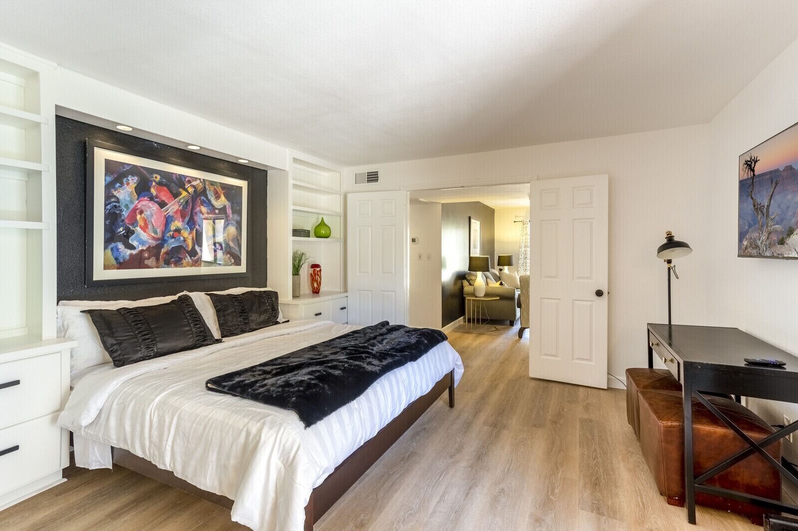 Un dormitorio con cama, escritorio y silla. Hay un cuadro en la pared y una puerta que da a otra habitación.