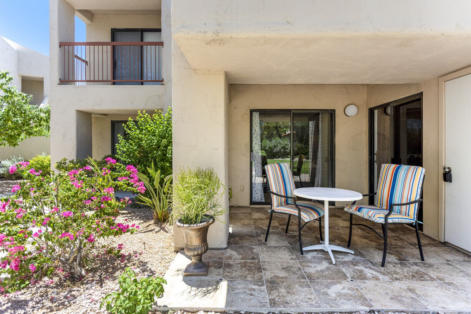 A patio with a round table and two striped chairs, surrounded by potted plants and flowers.