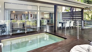 A modern outdoor pool area with glass railings, a wooden deck, and a dining area with white chairs. - Ripples on the Beach (Mission Beach)