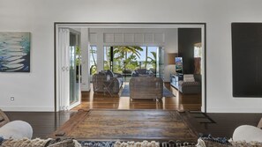 A modern living room with a large wooden table, wicker furniture, and a view of palm trees and the ocean through glass doors.
