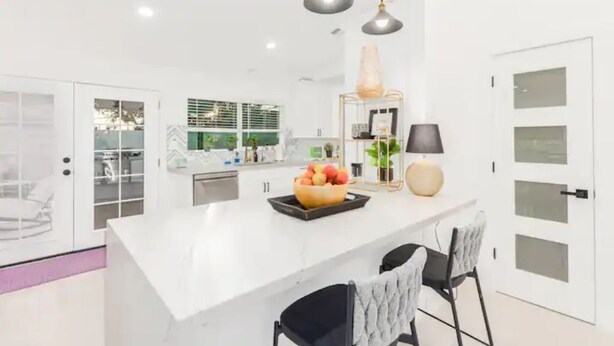 A modern kitchen with a white island, pendant lights, and a bowl of fruit.