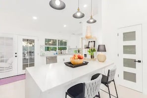 Uma cozinha moderna com ilha branca, luminárias pendentes e uma tigela de frutas.