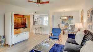Ein modernes Wohnzimmer mit Flachbildfernseher, einem blauen Sessel, einer Couch mit weißen Kissen, einem Couchtisch und einem Deckenventilator.