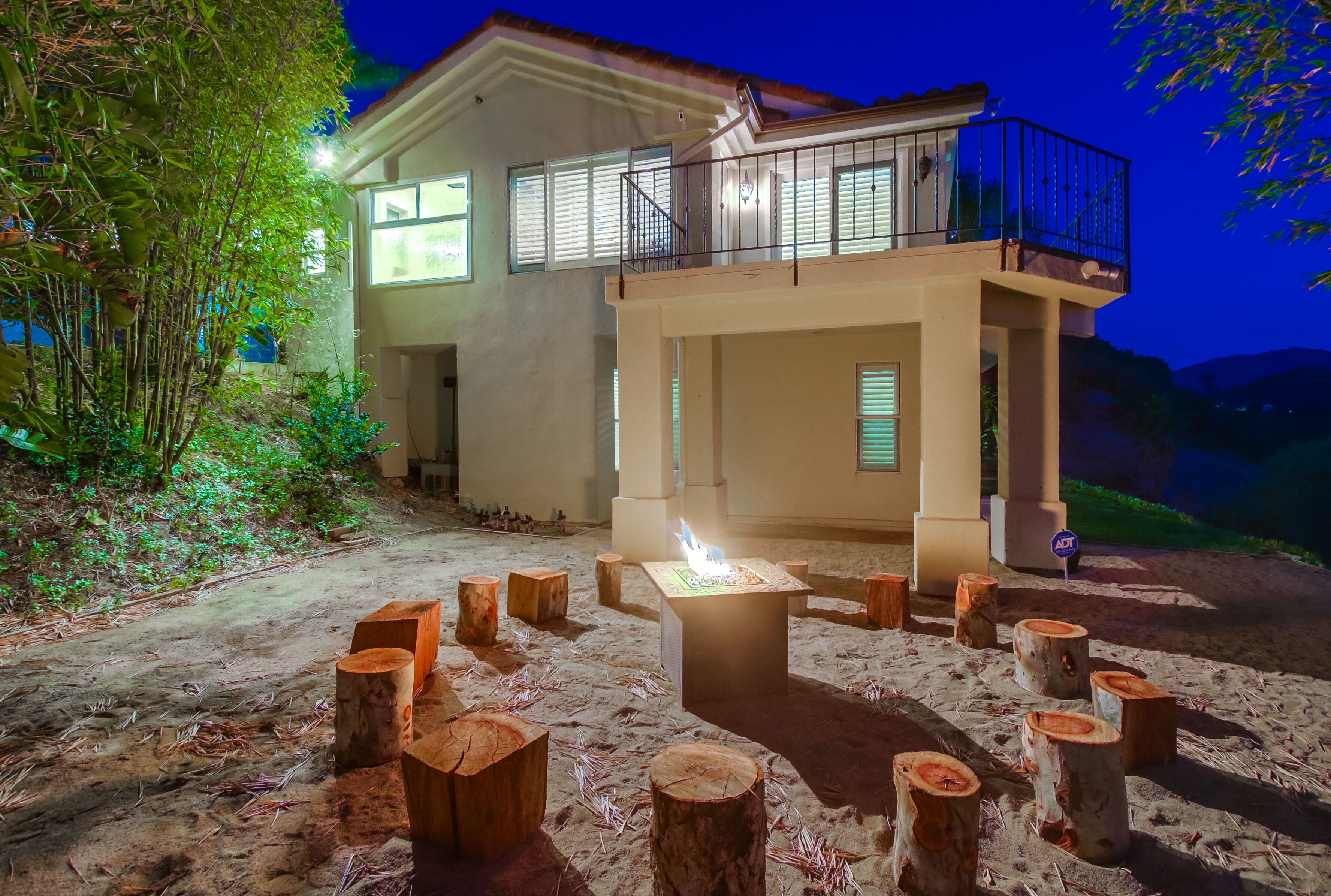 A two-story house with a balcony, surrounded by a sandy area with wooden stumps and a small table with a fire.