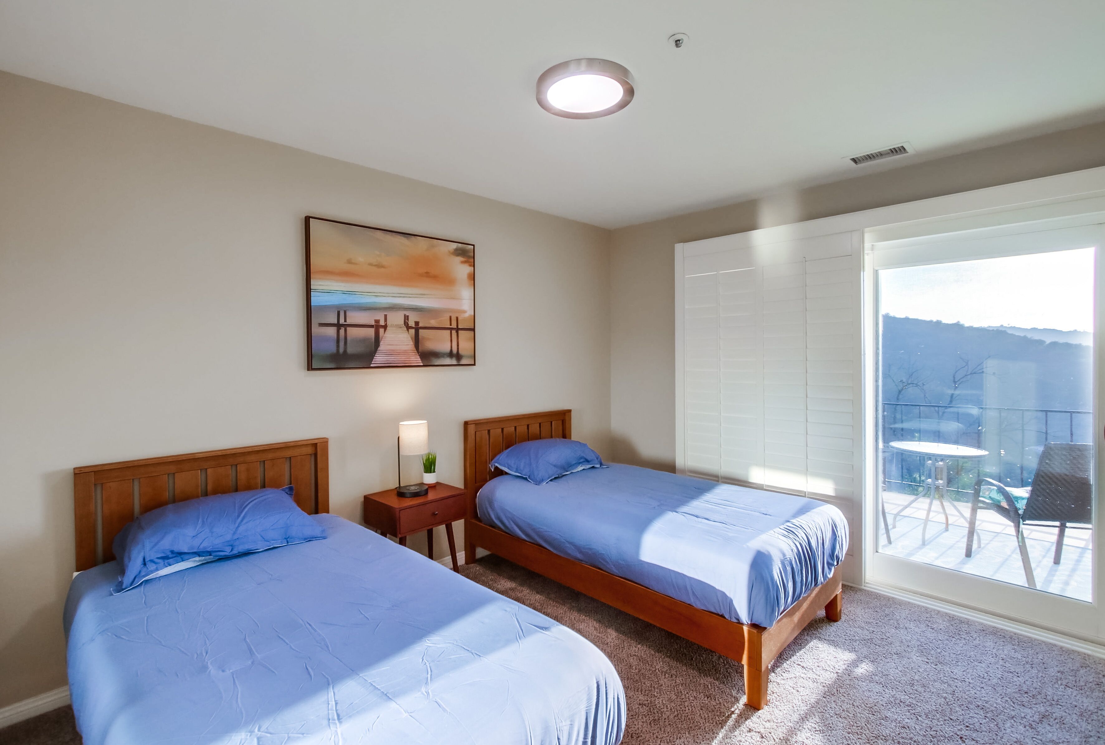 A hotel room with two beds, a painting on the wall, a nightstand, and a view of the outdoors through a large window.