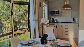 Ein Essbereich mit Tisch für vier Personen, eine Küche mit modernen Geräten und Blick auf einen Garten durch große Fenster.